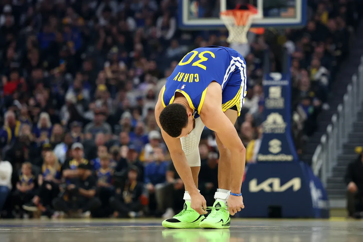 Steph Curry in a blue and yellow jersey bends down on the court to tie his bright green sneakers during a game, with a packed arena crowd in the background.