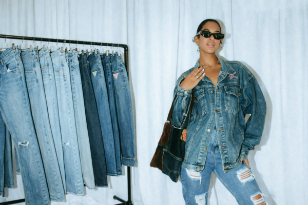 Model wearing Guess denim jacket and jeans standing next to a rack of blue jeans in a studio setting.