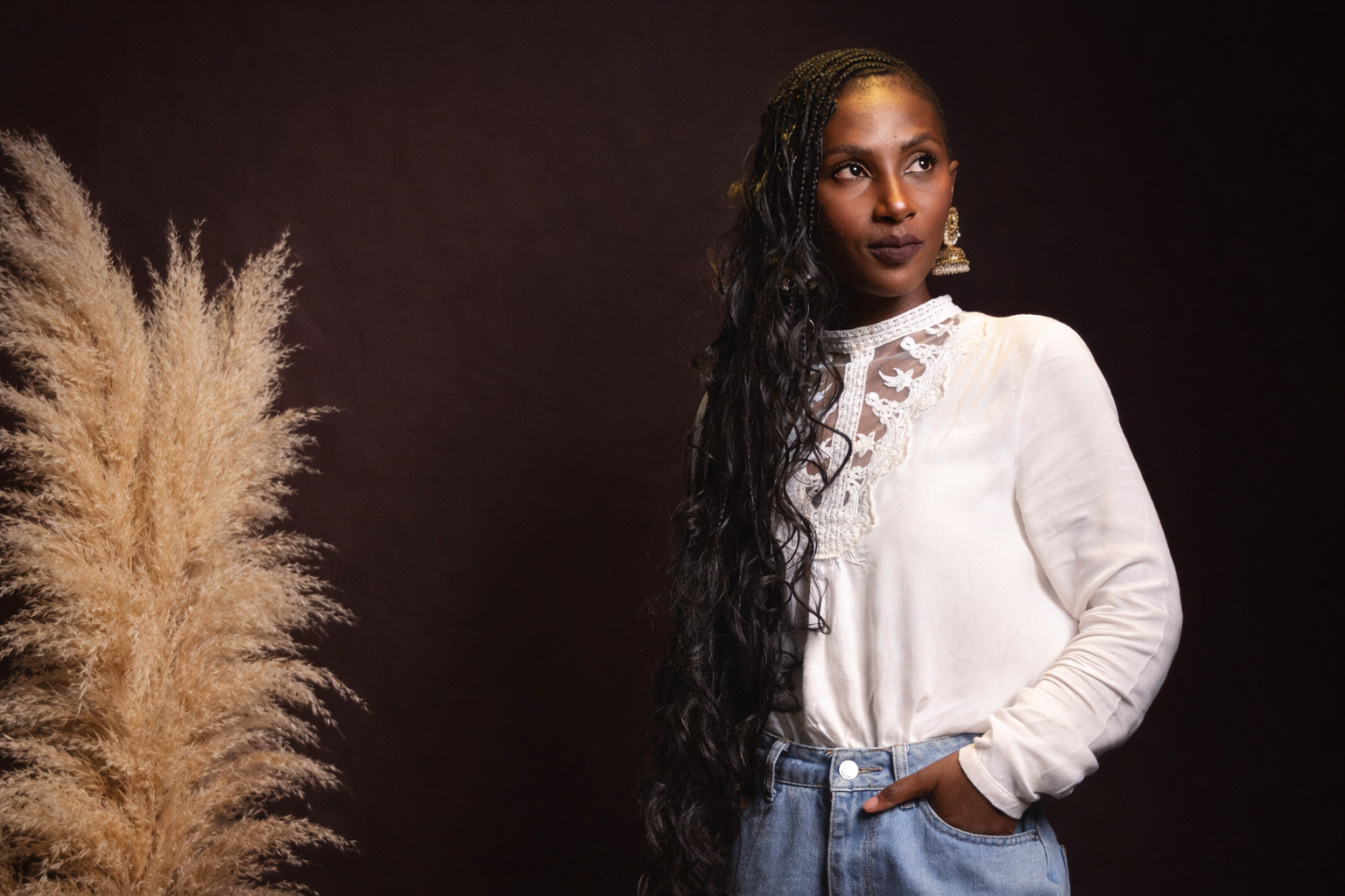 Portrait of Tamia wearing a white lace blouse and denim jeans, standing against a warm brown background with pampas grass, looking to the side.