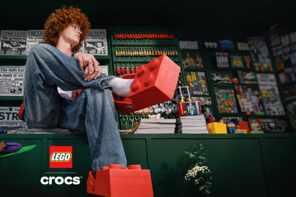 Model wearing oversized red LEGO-inspired Crocs shaped like building bricks inside a LEGO-themed retail space filled with colorful memorabilia and displays.
